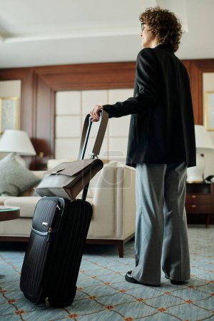 Foto de Joven mujer de negocios morena en pantalones grises y chaqueta negra de pie en la habitación de hotel y sosteniendo la maleta con su equipaje por asa - Imagen libre de derechos