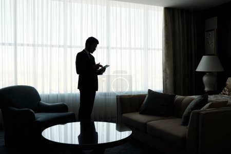 Foto de Esquema de hombre de negocios joven usando tableta mientras está de pie contra una gran ventana cubierta con cortinas de gasa blanca en la habitación de hotel - Imagen libre de derechos