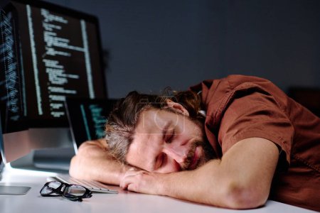 Foto de Joven programador masculino agotado con la cabeza en el escritorio por el teclado de la computadora durmiendo después del trabajo sobre el desarrollo de nuevo software de decodificación - Imagen libre de derechos