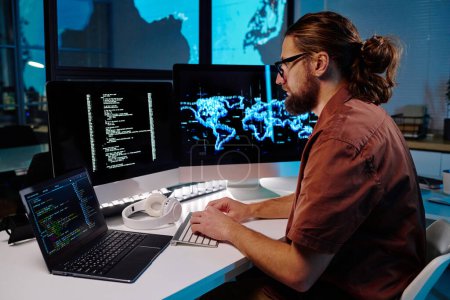 Foto de Joven programador masculino serio mirando datos codificados en la pantalla de la computadora mientras está sentado en el lugar de trabajo en la oficina oscura y trabajando por la noche - Imagen libre de derechos