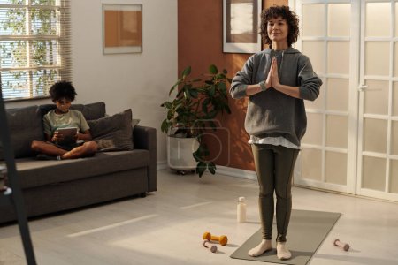 Young smiling woman practicing yoga exercise while standing on mat on the floor of living room while her little son relaxing on couch