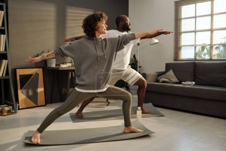 Foto de Mujer morena joven y su marido afroamericano de pie sobre esteras y haciendo ejercicio de estiramiento durante el entrenamiento en la sala de estar - Imagen libre de derechos