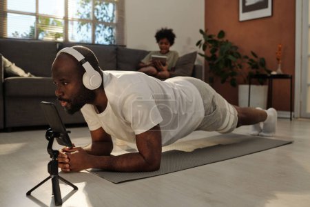 Photo for Young man in headphones and activewear repeating plank after fitness trainer on smartphone screen while exercising on mat at home - Royalty Free Image