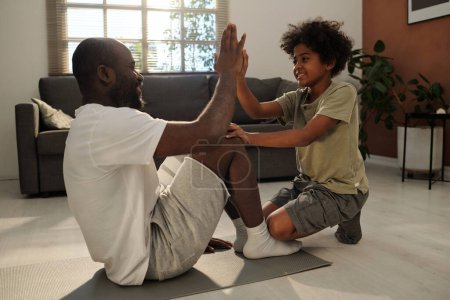 Photo for Happy little boy giving high five to his father in activewear sitting on mat during or after workout while spending weekend at home together - Royalty Free Image