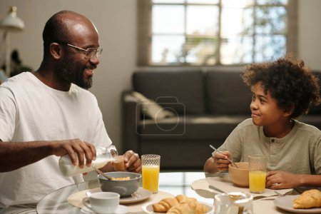 Photo for Young African American man in eyeglasses pouring milk in bowl with muesli and talking to his cute son while having breakfast with him - Royalty Free Image