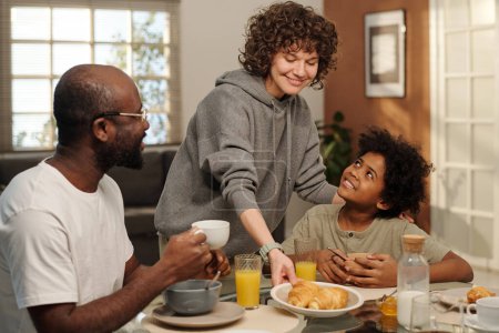 Téléchargez les photos : Jeune femme mettant assiette avec pâtisserie maison fraîche sur la table servi pour le petit déjeuner tandis que son mari et son fils ayant muesli avec du lait - en image libre de droit