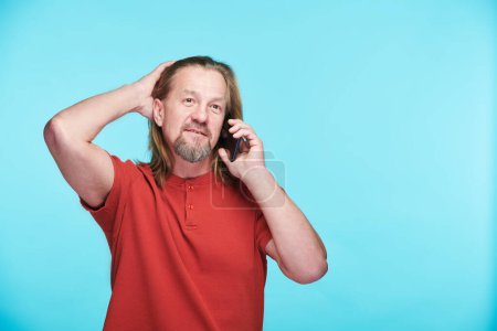 Photo for Mature man in red shirt having conversation on his mobile phone standing against blue background - Royalty Free Image