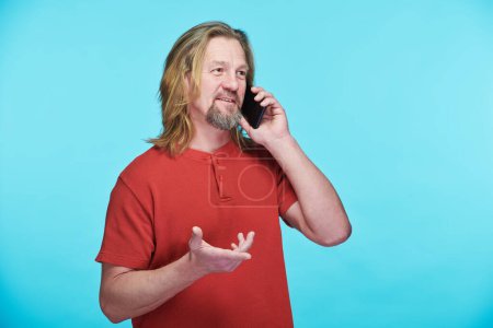 Foto de Retrato de hombre maduro con el pelo largo hablando en el teléfono móvil de pie sobre fondo azul - Imagen libre de derechos