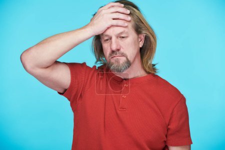 Photo for Mature bearded man in red T-shirt holding his head having depression standing against blue background - Royalty Free Image
