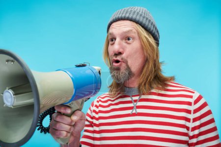 Photo for Mature man in striped shirt shouting in bullhorn to announce information standing on blue background - Royalty Free Image