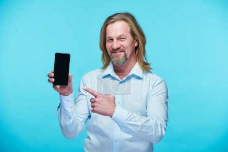 Téléchargez les photos : Portrait d'homme d'affaires mature en chemise pointant vers l'écran du smartphone et souriant à la caméra sur fond bleu - en image libre de droit