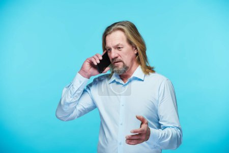Foto de Confiado hombre de negocios maduro que tiene una conversación seria en el teléfono móvil de pie sobre fondo azul - Imagen libre de derechos