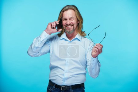Photo for Mature businessman with long hair talking on mobile phone and smiling at camera while standing on blue background - Royalty Free Image