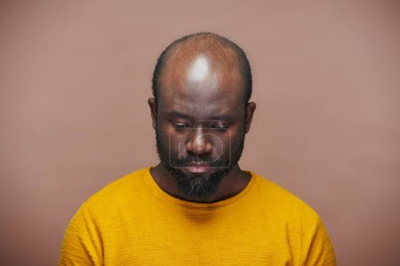 Foto de Retrato del hombre afroamericano en camisa amarilla mirando hacia abajo con expresión triste de pie sobre fondo marrón - Imagen libre de derechos