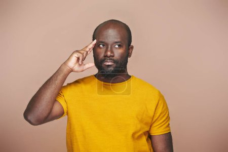 Photo for Portrait of African American pensive man in yellow shirt pointing at his head standing on brown background - Royalty Free Image