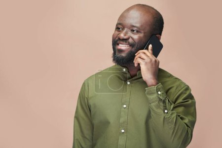 Photo for Smiling African American man in green shirt having conversation on mobile phone isolated on brown background - Royalty Free Image