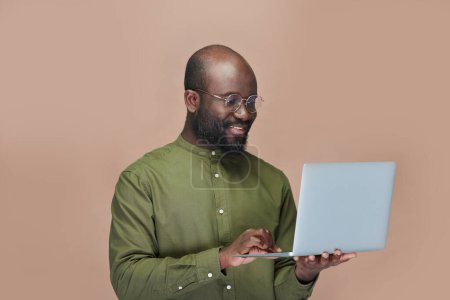 Photo for Smiling African American man holding laptop computer and typing on keyboard while standing on brown background - Royalty Free Image