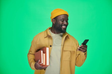 Téléchargez les photos : Afro-Américain avec des livres de lecture message sur son téléphone portable et souriant tout en se tenant debout sur fond vert - en image libre de droit