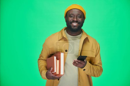 Foto de Retrato del hombre afroamericano con libros sonriendo a la cámara mientras usa su teléfono inteligente sobre fondo verde - Imagen libre de derechos