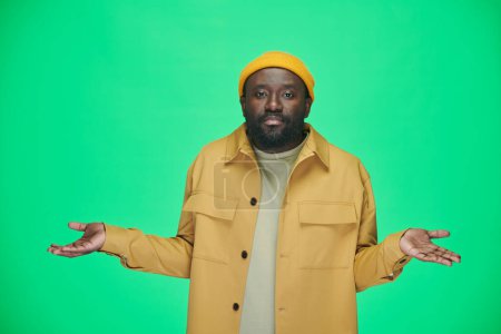 Téléchargez les photos : Portrait de l'homme afro-américain écartant ses mains dans le doute en regardant la caméra tout en se tenant sur fond vert - en image libre de droit