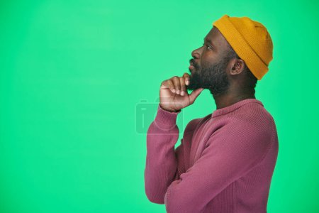 Téléchargez les photos : Vue latérale de l'homme afro-américain en chapeau jaune regardant loin avec expression coûteuse sur fond vert - en image libre de droit
