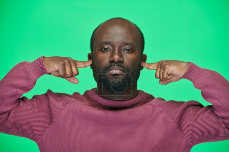 Téléchargez les photos : Portrait de l'homme afro-américain couvrant ses oreilles d'un bruit fort en regardant la caméra sur fond vert - en image libre de droit