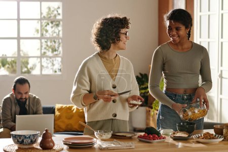 Téléchargez les photos : Jeune mère parlant et souriant avec sa fille adoptive pendant qu'ils préparent le petit déjeuner ensemble dans la cuisine - en image libre de droit