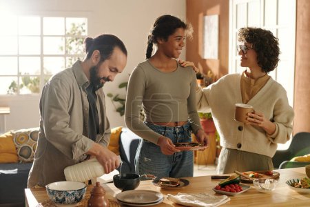 Téléchargez les photos : Famille de trois avec fille adoptive parlant entre eux et prenant le petit déjeuner ensemble dans la cuisine à la maison - en image libre de droit