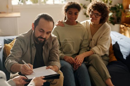 Photo for Dad signing custody contract during meeting with social worker with mom and adopted daughter sitting in background - Royalty Free Image
