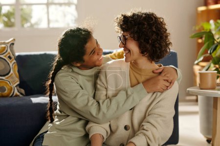 Téléchargez les photos : Afro-Américaine heureuse adolescente embrassant sa mère adoptive pendant qu'ils passent du temps ensemble à la maison - en image libre de droit