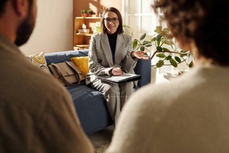 Photo for Young social worker in suit sitting on sofa in the living room and talking to foster parents about custody at meeting at home - Royalty Free Image