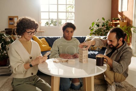Photo for Father playing board game together with his adopted daughter while mother using her smartphone, they sitting in the living room at home - Royalty Free Image