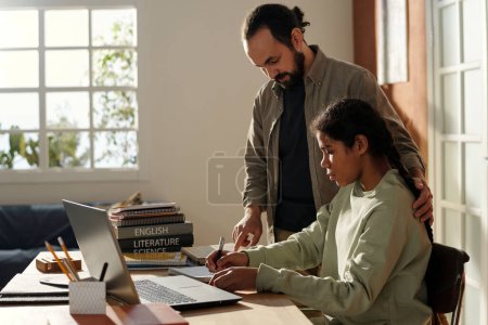 Foto de Joven padre ayudando a su hija con su tarea mientras ella trabaja con el ordenador portátil y libros en la mesa - Imagen libre de derechos