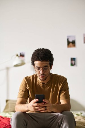 Photo for Vertical image of young man in eyeglasses reading message on smartphone with sad expression while sitting alone in his bedroom - Royalty Free Image