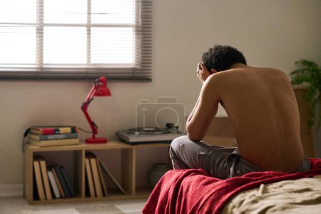Téléchargez les photos : Vue arrière du jeune homme en dépression assis seul dans sa chambre et inquiet de ses problèmes - en image libre de droit