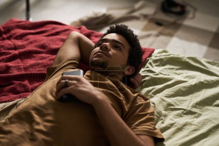 Téléchargez les photos : Vue grand angle de gars couché sur le lit avec smartphone et penser à quelque chose - en image libre de droit