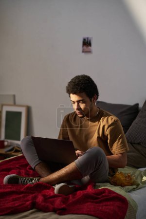 Foto de Imagen vertical del adolescente usando portátil mientras está sentado en su cama y comiendo papas fritas - Imagen libre de derechos