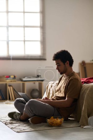 Foto de Imagen vertical del joven sentado en el suelo comiendo patatas fritas y comunicándose en línea usando su portátil - Imagen libre de derechos