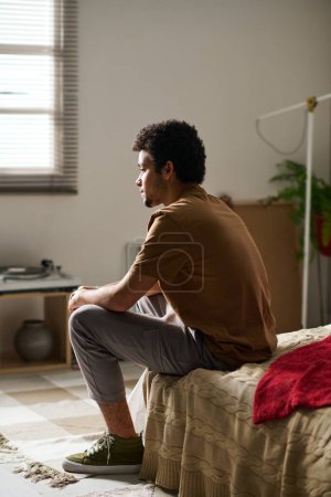 Téléchargez les photos : Vue arrière du jeune homme assis sur le lit dans les pensées tout en restant seul dans sa chambre - en image libre de droit