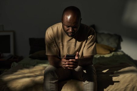 Foto de Hombre afroamericano leyendo el mensaje en el teléfono inteligente mientras está sentado en la cama en el dormitorio - Imagen libre de derechos