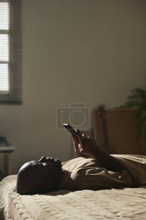 Téléchargez les photos : Image verticale d'un homme mélancolique afro-américain lisant un message sur son smartphone alors qu'il était allongé sur son lit dans sa chambre - en image libre de droit