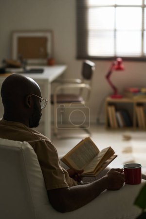 Téléchargez les photos : Image verticale de l'homme afro-américain appréciant la lecture assis sur un fauteuil avec une tasse de thé à la maison - en image libre de droit