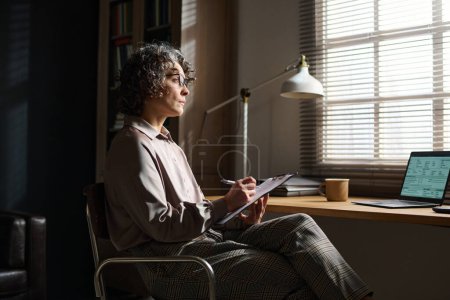 Photo for Side view of mature professional psychologist looking through venetian blinds on window while sitting by workplace and making notes - Royalty Free Image