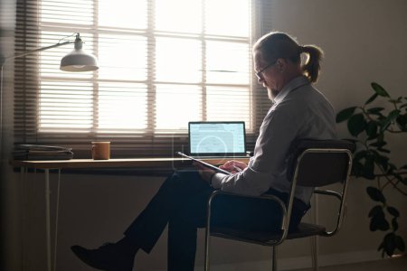 Foto de Psicólogo masculino maduro en ropa casual sentado en la silla por el lugar de trabajo en la oficina y mirando a través de documento con datos personales de los pacientes - Imagen libre de derechos