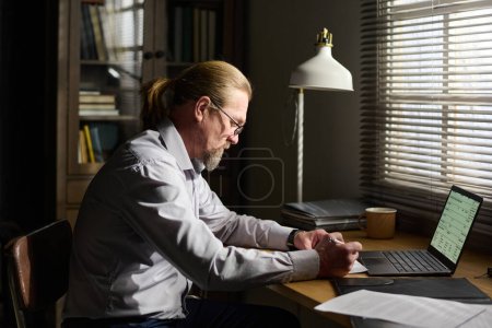 Foto de Psicoterapeuta varón maduro confiado que toma notas en documento mientras que se sienta por el escritorio delante de la ventana y se prepara para la nueva sesión - Imagen libre de derechos