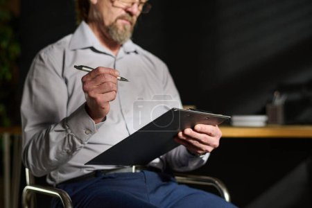 Foto de Primer plano del psicólogo masculino maduro sosteniendo el portapapeles y la pluma mientras está sentado en la silla delante de la cámara y tomando notas en el documento - Imagen libre de derechos