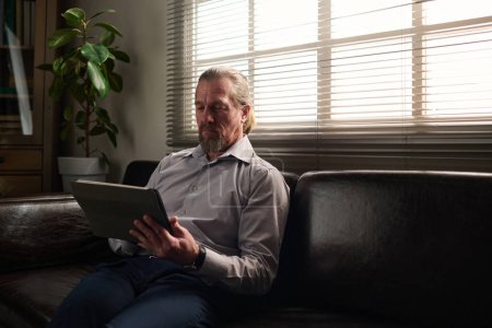 Foto de Psicólogo maduro confiado mirando la pantalla de la tableta durante la sesión en línea con el paciente mientras está sentado en el sofá de cuero negro por ventana - Imagen libre de derechos