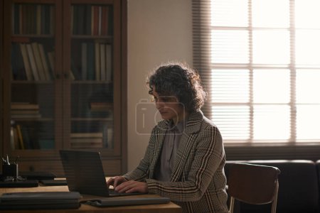 Foto de Consejera femenina madura grave escribiendo en el teclado de la computadora portátil mientras está sentada en el lugar de trabajo en la oficina y consultando a pacientes en línea en problemas - Imagen libre de derechos