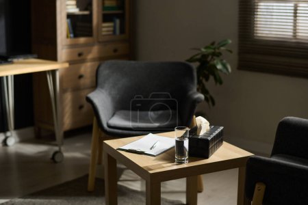 Téléchargez les photos : Partie du bureau du psychothérapeute avec petite table carrée en bois avec document, verre d'eau et boîte entourée de deux fauteuils - en image libre de droit