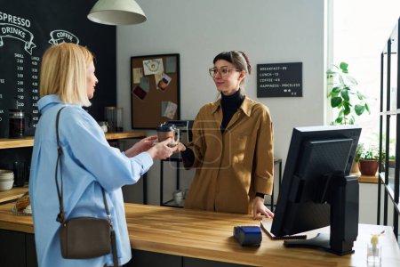 Photo for Young cafe clerk in casualwear passing two cups of coffee to mature female guest over counter desk while serving her in the morning - Royalty Free Image